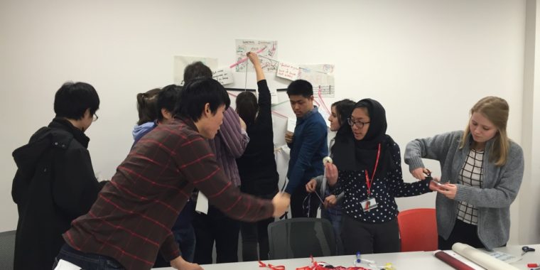 This image shows students crowded around a big table covered with card, paper, string, ribbon, glue, scissors and tape. Behind them they are creating a giant network map covering the whole wall of the classroom with their ideas about sustainability and how these ideas are linked together.
